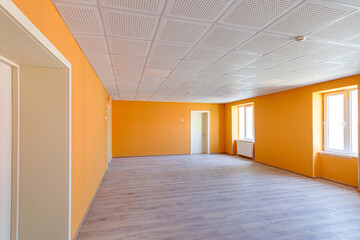 Obraz premium Spacious empty office or classroom interior featuring bright orange walls, wooden laminate flooring, white doors, and a professional acoustic grid ceiling with windows providing natural light.
