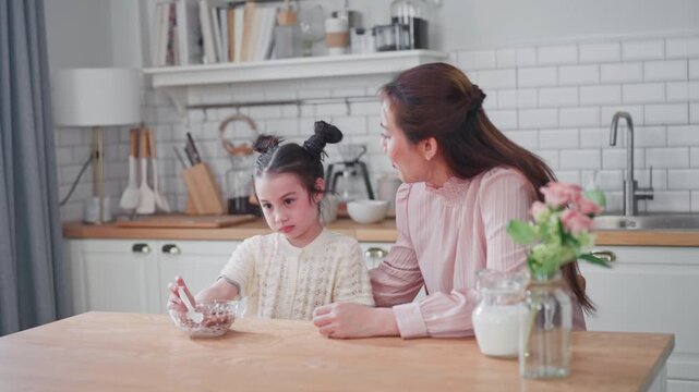 Loving mother takes care of her child during breakfast. The happy mother watches her daughter eating cereal together in their kitchen at home.
