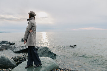 Woman in grey coat and hat stands on rocky shore looking at sea during cloudy day. Calm water and soft light create peaceful, contemplative mood by the coast. © SHOTPRIME STUDIO