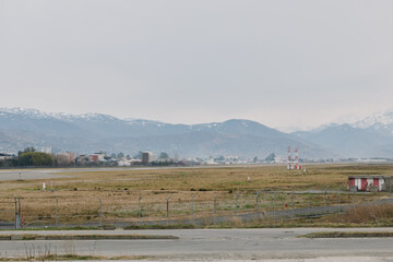Fototapeta premium Empty airfield with runway and small buildings in front of hazy mountains under cloudy sky. Concept for aviation, transportation, and industrial landscape with wide open space.
