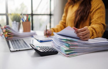 A woman in a mustard sweater organizes colorful paperwork on her desk, while using a laptop,...