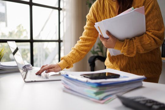 A woman in a yellow knit sweater multitasks at her desk, balancing paperwork and technology, embodying a productive and organized office environment.