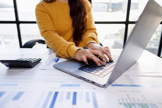 Close-up of a woman's hands on a laptop keyboard analyzing budget data, illustrating engagement with digital financial tools amidst spreadsheets and calculators.