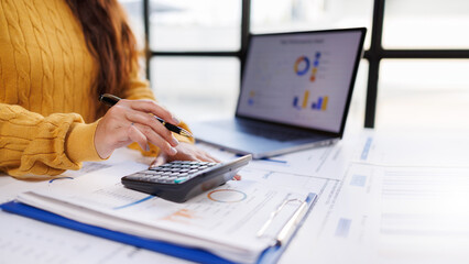 A woman in a cozy yellow sweater is engaged in calculating figures on a calculator while reviewing financial reports next to a laptop, embodying financial diligence. © groviapoto