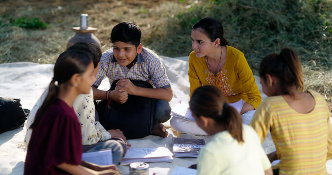 Indian villager girl boy sitting on mat do study read book outdoor rural area ground place. Happy group child hold notes talk gossip enjoy desi life new day open class field