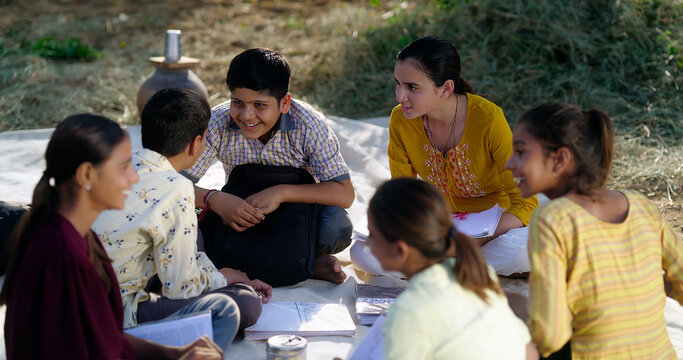 Indian villager girl boy sitting on mat do study read book outdoor rural area ground place. Happy group child hold notes talk gossip enjoy desi life new day open class field