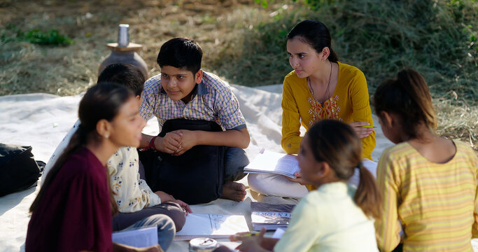 Indian villager girl boy sitting on mat do study read book outdoor rural area ground place. Happy group child hold notes talk gossip enjoy desi life new day open class field