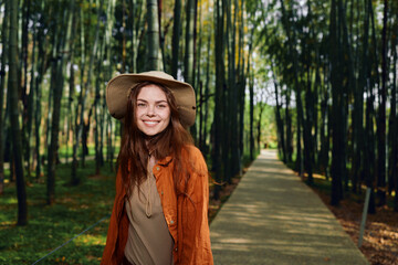 Fototapeta premium Woman smiling in a straw hat walking in an autumn park on a tree lined path, portrait of a happy young adult wearing a cozy jacket with natural light and seasonal foliage.