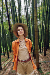 Fototapeta premium Woman in bamboo forest wearing straw hat and casual orange shirt, smiling portrait amid tall green stalks, nature walk in summer outfit, relaxed pose and soft natural light.