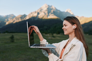 A cheerful woman works outdoors with a laptop in a scenic field beneath mountain peaks. She smiles...