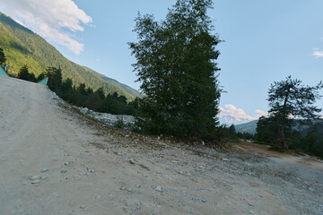 Fototapeta premium A rugged mountain road winds through a rocky landscape under a bright blue sky. Sparse trees line the track, creating a sense of solitude and outdoor exploration.