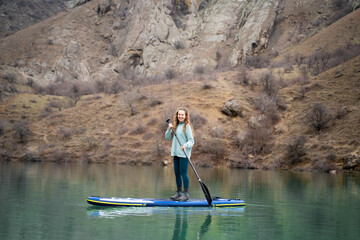 Naklejka premium Woman on paddle board on mountain lake in daylight. Young sporty female on SUP surf on water with mountains in the background. Wide scenic view.