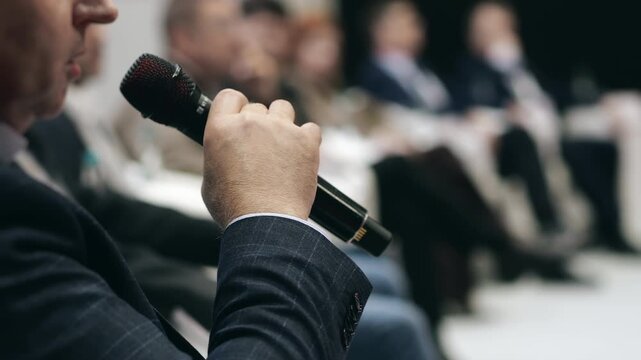 Man speaking into microphone at business conference panel discussion, audience participation and professional communication at corporate event indoors.