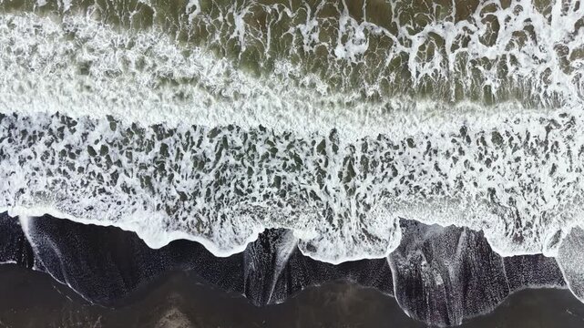 Waves roll onto a beach, breaking against the dark sand and rocky shore. White foam forms as the water recedes. The scene shows the shoreline under bright daylight.