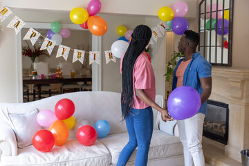 Couple holding colorful balloons and standing in living room under Happy Birthday banner