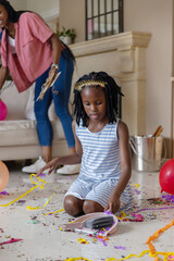 African American mom and child kneeling, picking confetti with dustpan in living room, balloons