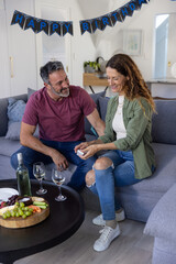 Senior couple sitting on gray sectional sofa in living room, holding white gift box beside wine