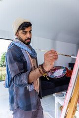 Asian man wearing knit beanie painting on stretched canvas in home studio holding palette and brush