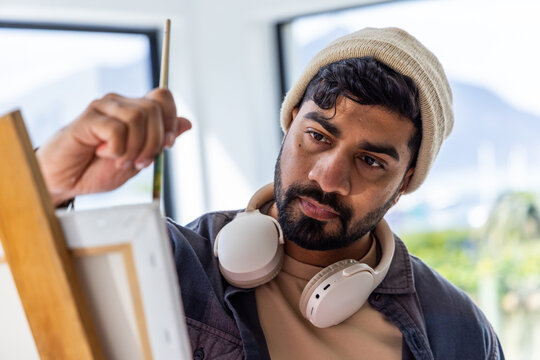 Indian man painting on canvas on easel in studio holding brush, wearing beanie and headphones