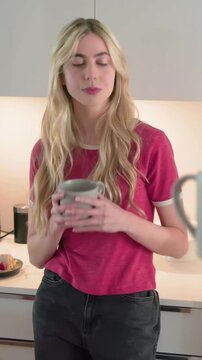 Vertical video: Talking lady in red tee with gray mug in kitchen as friend coming with yellow strap