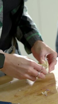 Vertical video: Grabbing garlic bulb, plaid woman peeling clove at counter pal slicing for cooking