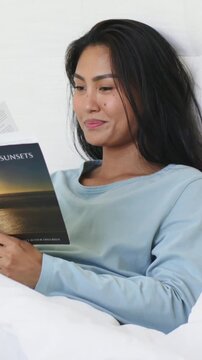 Vertical video: Reading Asian woman holding SUNSETS book in bed, turning page and smiling at pages