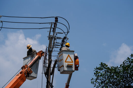 Two electrical workers repairing power lines on utility pole, wearing safety helmets and protective gear, performing maintenance on high voltage distribution system against blue sky background