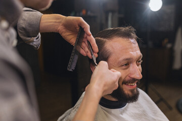 Barber cutting male client hair with scissors and comb. Close up of professional haircut process, precision technique, men grooming service in modern barbershop.