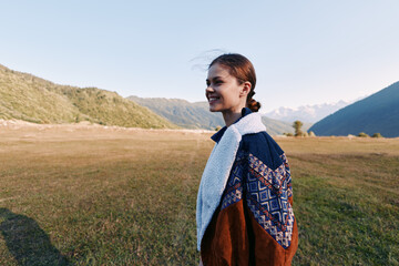 Naklejka premium Woman, smile, landscape, mountains, meadow, sweater, outdoors, portrait — Young woman smiling in a cozy sweater standing in a meadow by mountain landscape at golden hour for travel and nature.