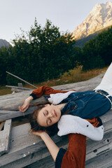 Naklejka premium Woman portrait relaxing on wooden deck in nature with mountains in background, eyes closed and cozy layered denim and shearling jacket, peaceful outdoor leisure moment and soft evening light