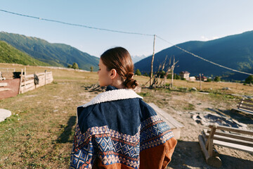 Naklejka premium Girl in patterned sweater back view in rural village landscape with wooden fences and distant mountains, outdoor portrait of youth exploring countryside pathway under clear blue sunlight.