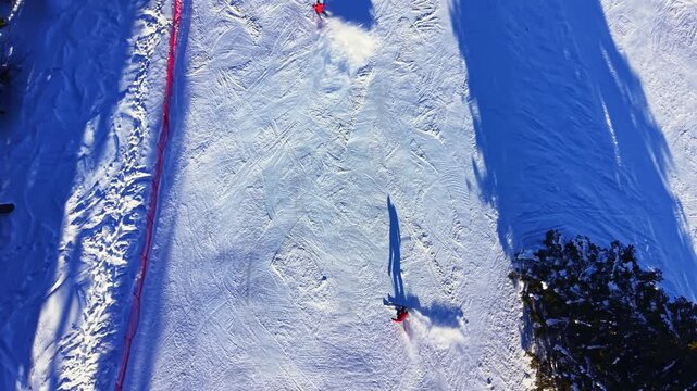 Two skiers navigate a snowy slope at a ski resort, leaving trails in the freshly fallen snow. Sunlight casts long shadows on the terrain, creating a bright scene.