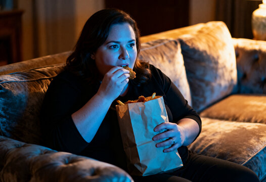 Woman eating unhealthy fast food while sitting on sofa at night. Overeating and comfort food habit. Concept of unhealthy lifestyle, emotional hunger and sedentary behavior at home.