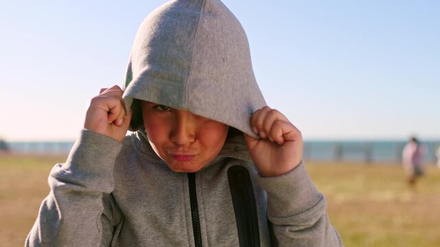 Beach, funny face and hoodie with boy child outdoor in summer for travel, trip or vacation. Crazy, humor and squint with silly kid on grass lawn at coast for comedy, goofy or playful expression
