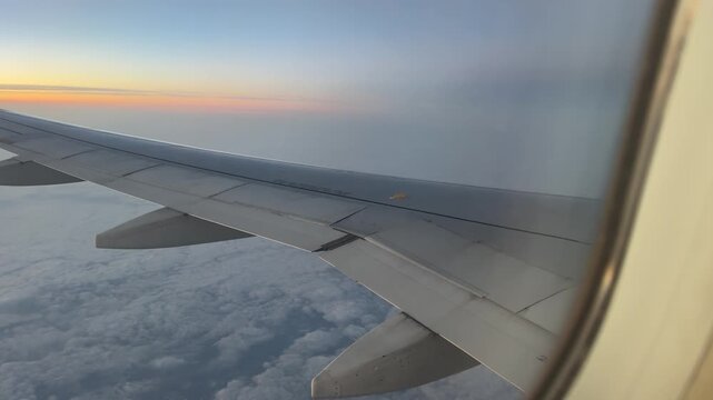 Passenger&rsquo;s view from airplane window of the wing glowing in the warm sunset light, with clouds below and vibrant colors painting the sky in an unforgettable scene