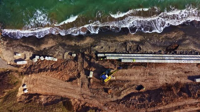 Workers use heavy machinery to build structures on the beach. The shore is seen with equipment beside the water. Construction materials are placed along the coastline.