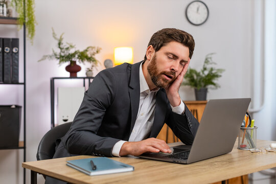 Middle-aged businessman at home office types report on laptop gets sleepy and nods off leaning on hand. Freelancer guy at table fights fatigue blinks slow and almost sleeps then returns to work again.