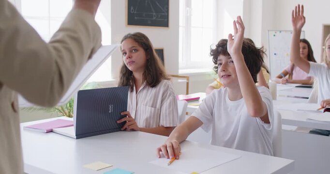 Teenager schoolboy sitting at the desk in the classroom raising hand up and giving the right answer to female school teacher. Back to school and education concept. 4k video. Slow motion video.