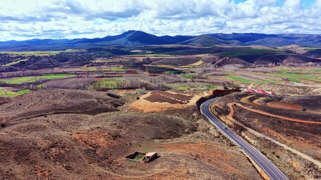 Green and brown fields along with a winding road surrounded by hills. The sky is bright with clouds, and some buildings are visible in the distance.