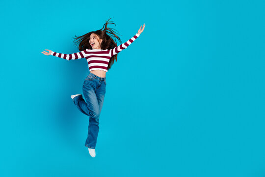 Happy young woman jumping with joy on a bright blue background wearing striped sweater and jeans &mdash; energetic casual lifestyle portrait expressing freedom and youthful fun