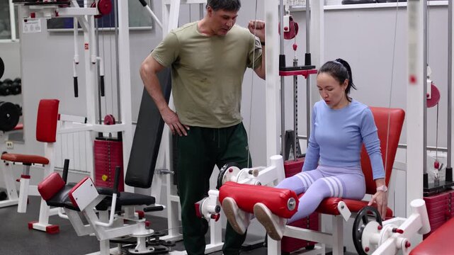 Slow motion 4k footage of a middle-aged woman during gym workout. Leg extensions under the supervision of a trainer. Leg extensions in gym fitness exercise.