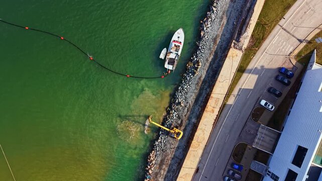 A yellow machine is working on the shoreline to clean the water while a boat is anchored nearby. Cars are parked along the road next to the water.