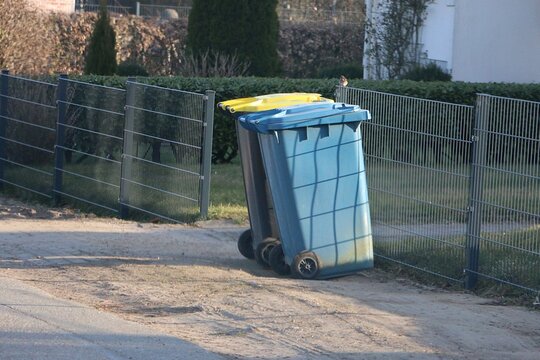 Two differently colored garbage cans, one blue and one dark with a yellow lid, stand on the street at a metallic fence

