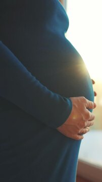 A pregnant woman gently holds her round belly while wearing a fitted dress. The woman stands near a window, showcasing the contours of her pregnancy in a soft light.