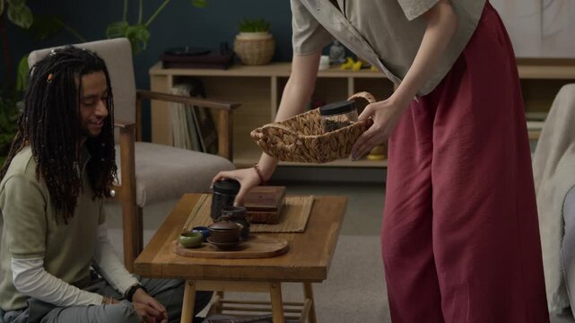 Handheld shot of young Asian woman setting table with cups for home tea ceremony at wooden table with multiethnic boyfriend