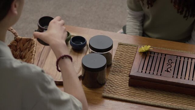 High angle view of young Asian woman serving tea canisters and cups for traditional tea ceremony with boyfriend sitting at wooden table