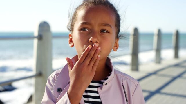 Girl, wave and face with kiss at beach on promenade, outdoor and happy on vacation in summer. Child, smile and playful with fun, hello or hand gesture for portrait on seaside boardwalk in Colombia