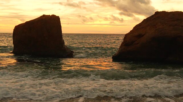 Waves, sunset, seascape washing onto beach between two large sea cliffs during beautiful golden hour.