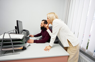 A blonde woman leans over a man seated at a computer, offering guidance in a professional office...
