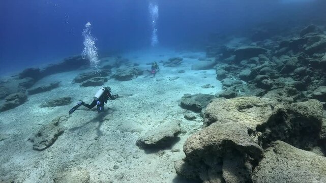 Slowly swimming pair of scuba divers exploring underwater landscape in the sea. Sand and rocks, clear ocean and swimming underwater explorers. Divers in the seascape, vivid blue ocean.